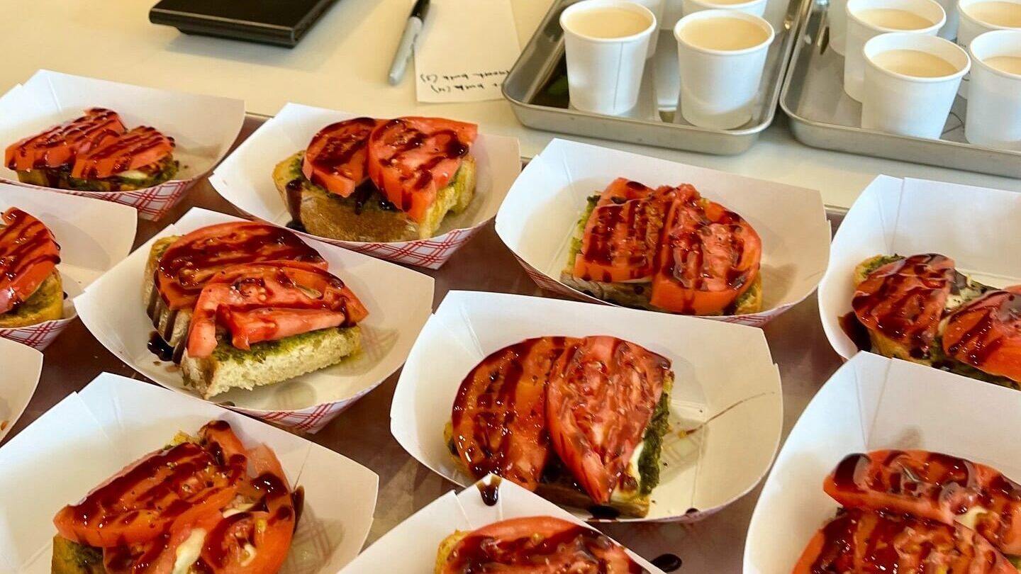 slices of bread topped with fresh tomato slices, a green spread (likely pesto), and drizzled with a dark sauce, arranged neatly in paper trays with cups in the background